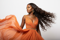 brown skin african american woman in glamours orange attire modeling long body wave hair, head turned to the side to showcase the hair, white studio background, natural glam makeup, gold accessories